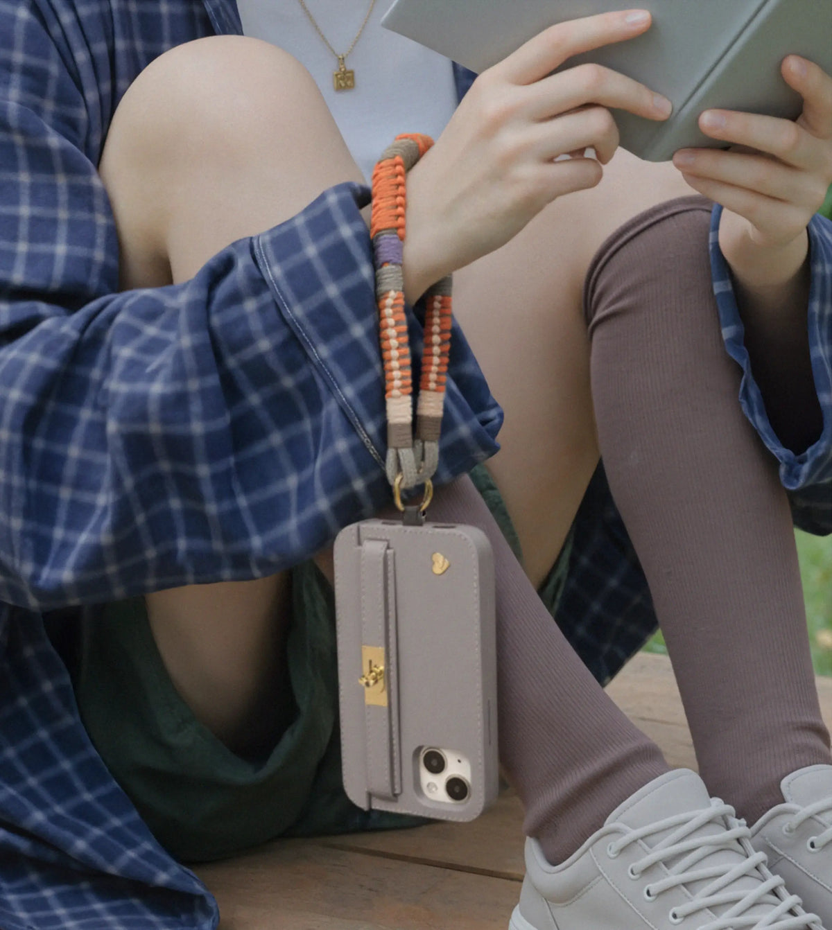 Woman reading in garden with Amber Haze HAND-BRAIDED TENCEL™ phone strap suspending Elephant gray LUMI iPhone 15 case. HAND-BRAIDED TENCEL™ WRISTLET.