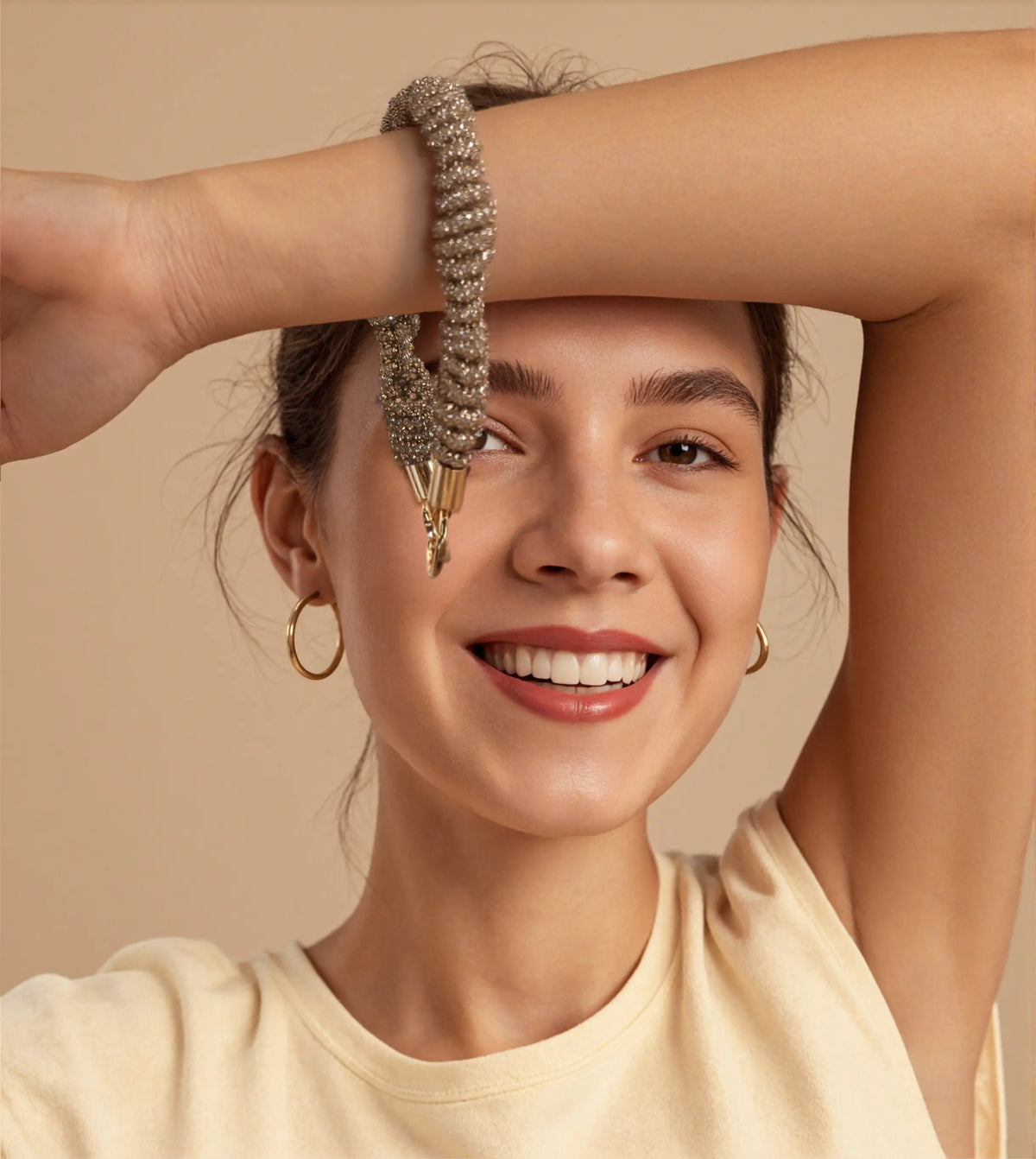 Joyful woman with gold earrings raising hand to display HAND-BRAIDED CRYSTAL WRISTLET with clasped buckles. OTHER STRAPS for elevated moments.