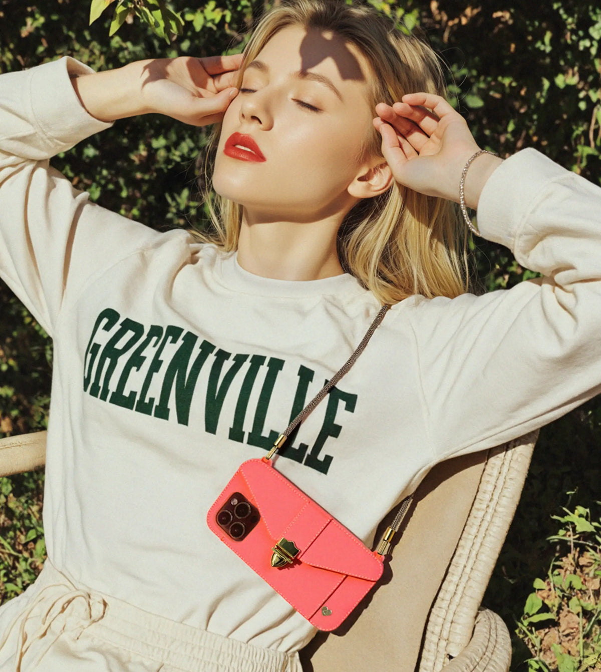 Sunlit relaxation: Blonde woman in garden with golden Bling Bling wristlet suspending Tango orange AURA iPhone case. IPHONE PURSE CASES for serene moments.